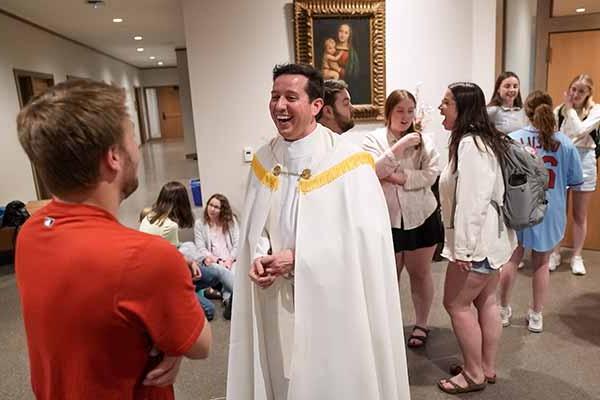 A man wearing white vestments speaks to a student. Other students can be seen chatting in the background.