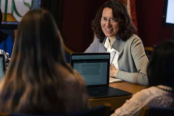 A female faculty member speaks with students while sitting around a table. The students have laptops open to work on.